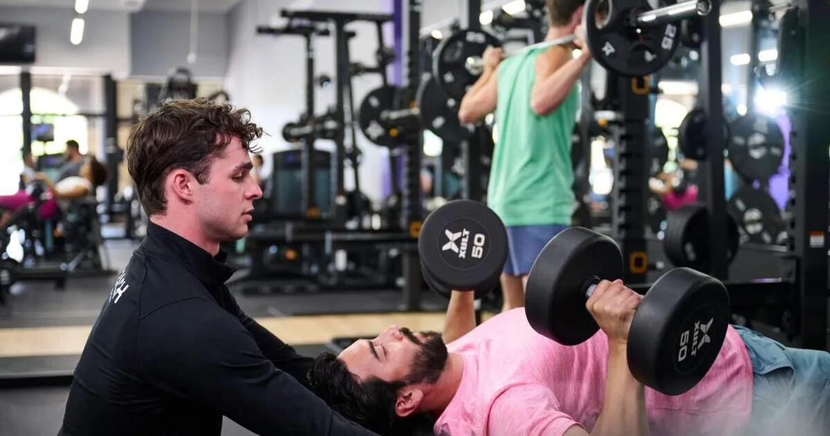 Personal trainer spotting client doing dumbbell bench press in busy gym