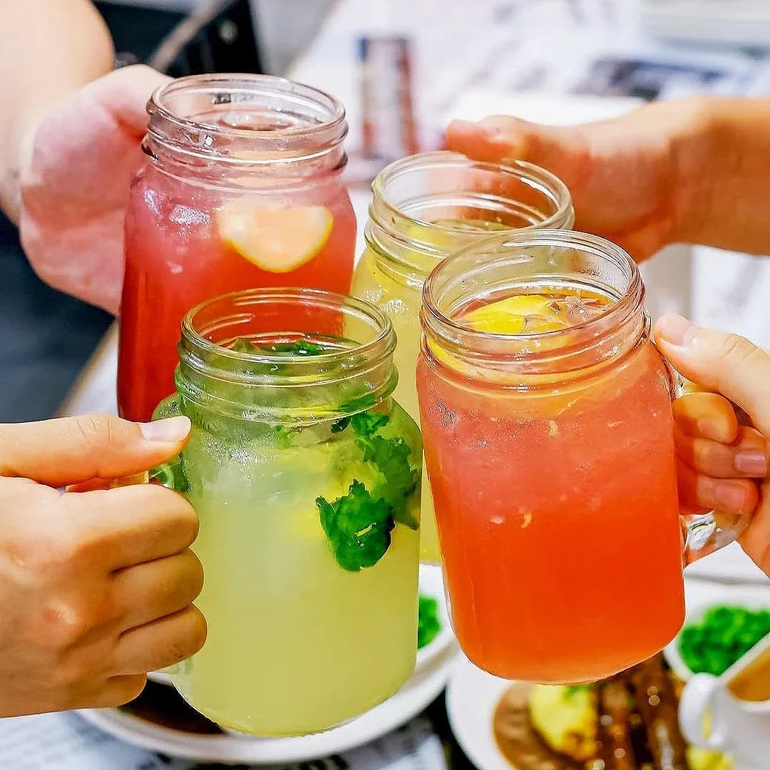 Three chilled drinks in mason jars: green mojito, orange and red beverages held by diners