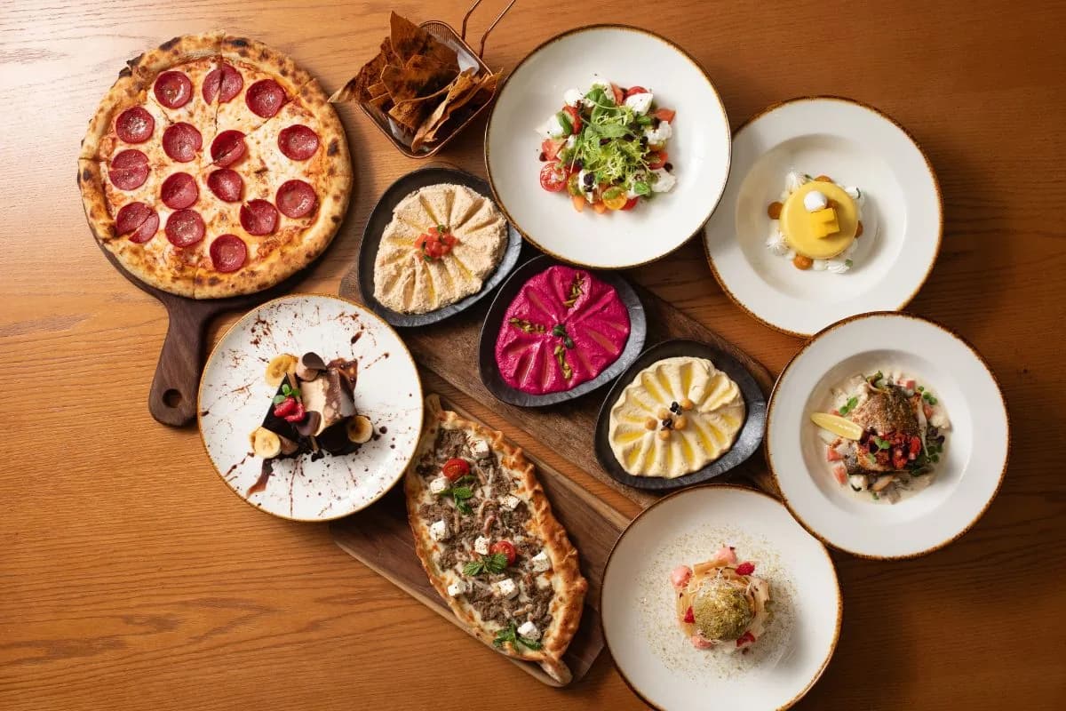 Flatlay of restaurant dishes: pepperoni pizza, plated salads, appetizers, and dips on wooden table