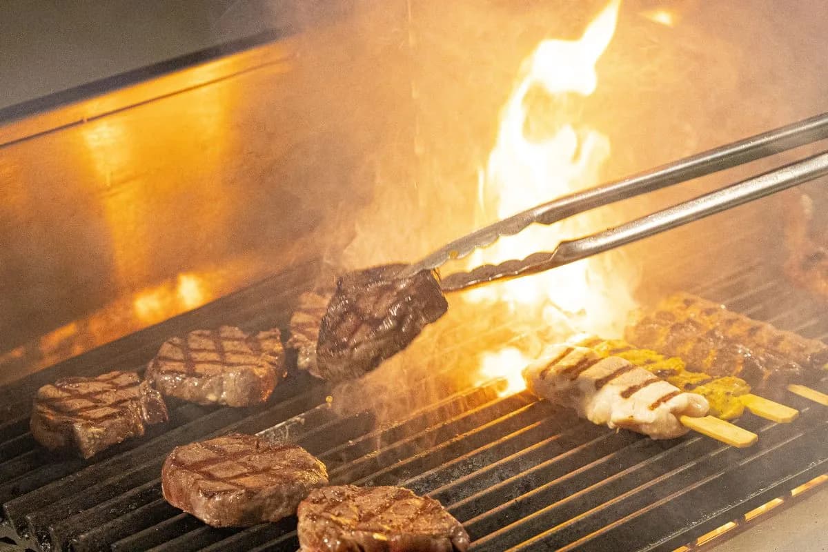Steaks and fries cooking on a grill with open flames and tongs holding meat