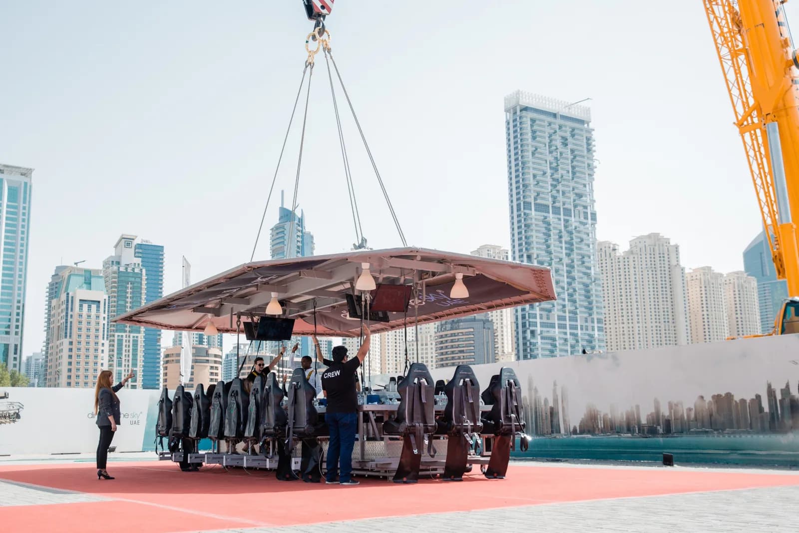 Suspended dining platform with guests and crew being hoisted by crane above a waterfront with city skyline