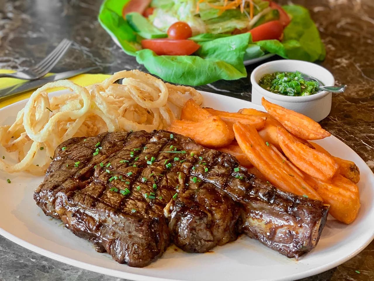 Grilled steak with onion rings, sweet potato fries, and fresh salad
