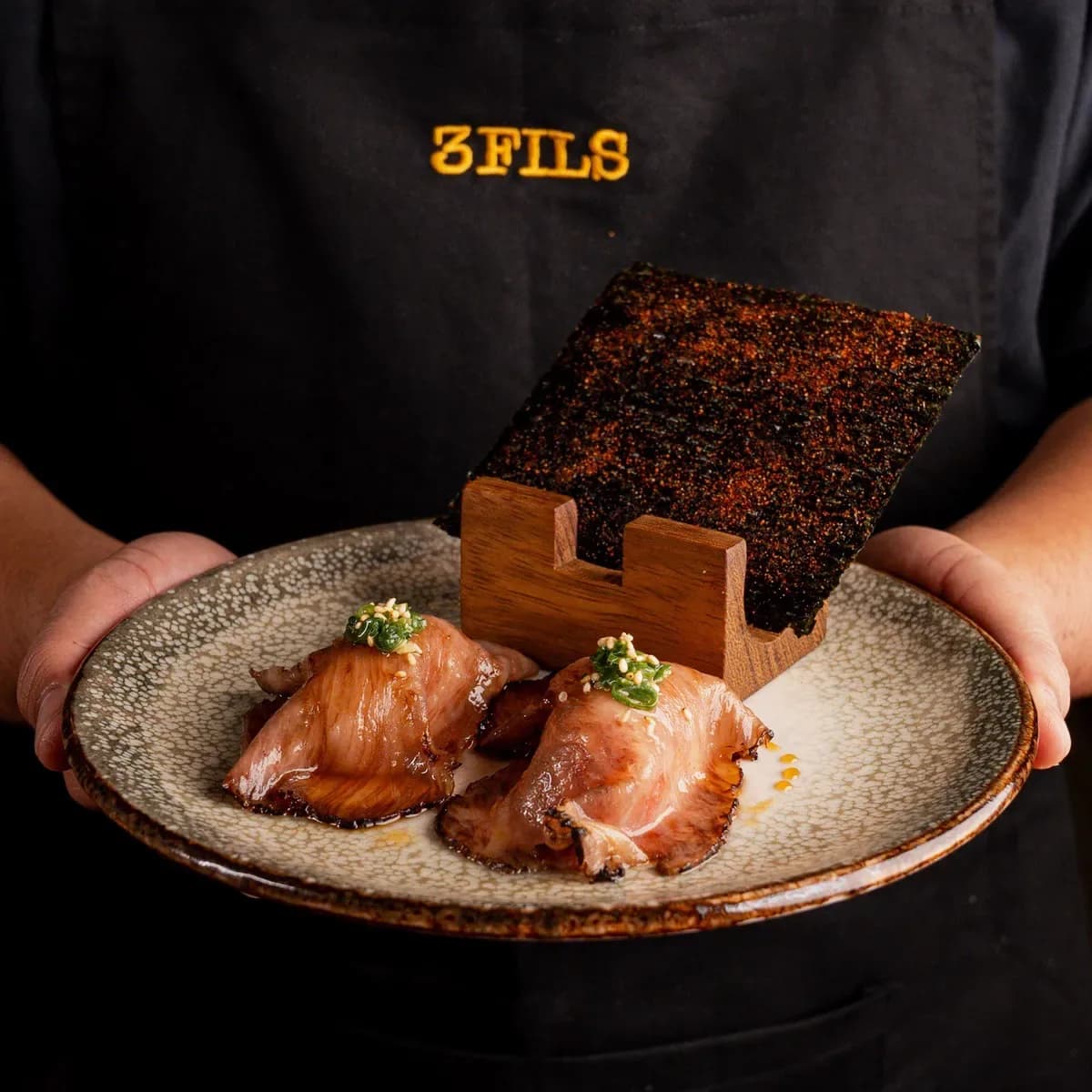Server holding a ceramic plate with seared fish, microgreens, and sauce, with nori crisp in wooden holder