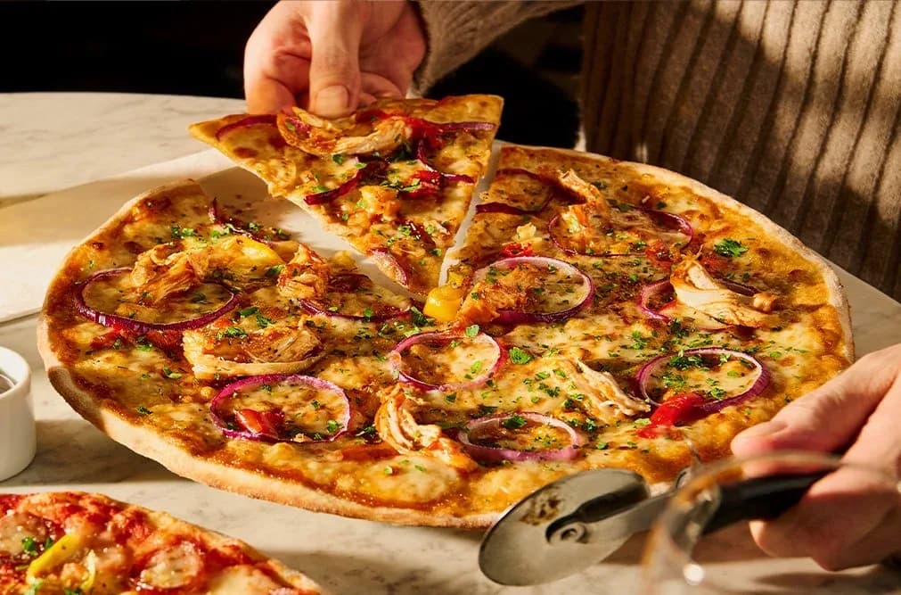 Hand lifting a slice of pizza topped with cheese, red onions, and herbs from a whole pizza