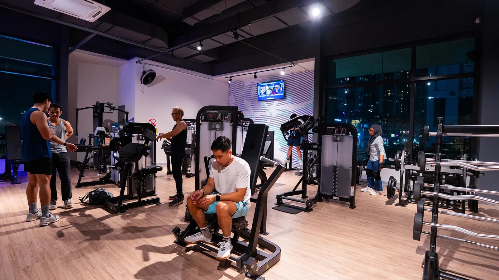 Gym members exercising on various machines in a bright, modern fitness facility with city views