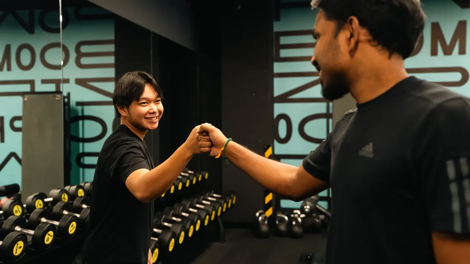 Trainer and client fist bump at gym with dumbbells and signage behind them