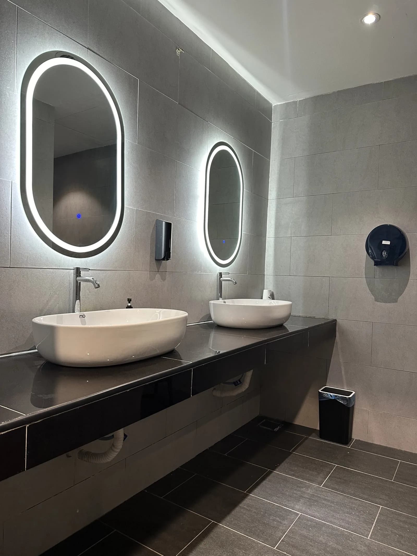 Modern gym bathroom with dual white sinks, LED-lit mirrors, and gray tile walls