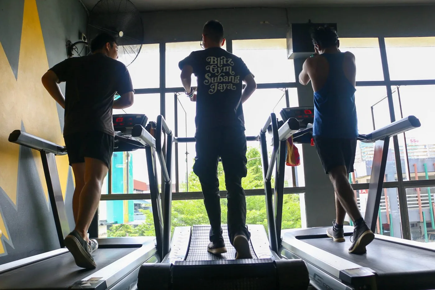 Three gym members running on treadmills with city views through large windows