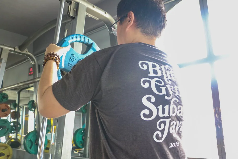 Person in Blitz Gym shirt holding blue climbing carabiner in gym space with equipment