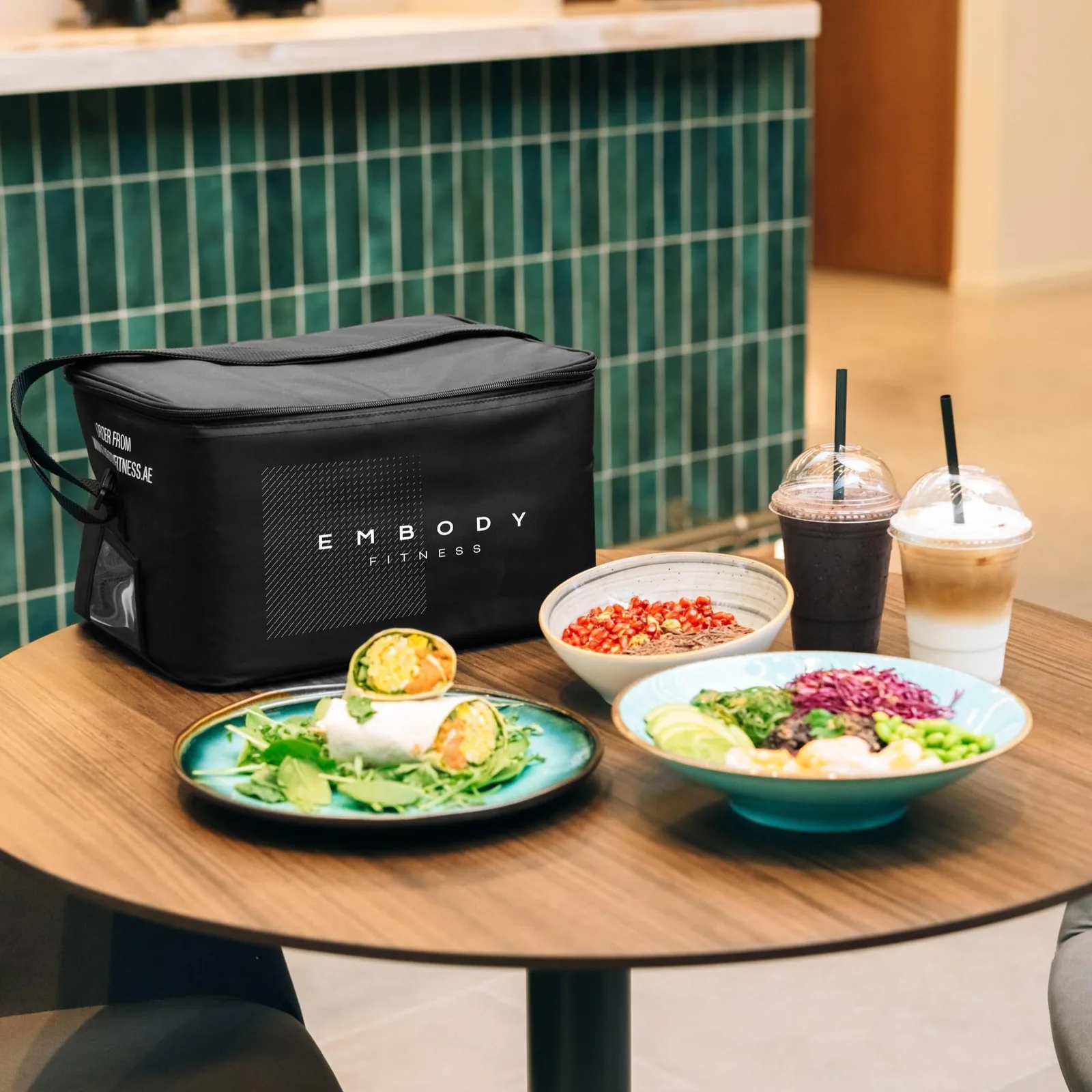 Healthy meal prep plates with vegetables, eggs, and beverages next to an Embody Fitness branded cooler bag
