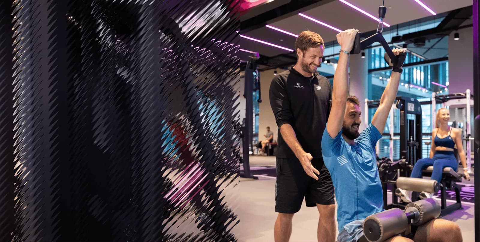 Personal trainer assisting client with overhead dumbbell exercise in bright modern gym