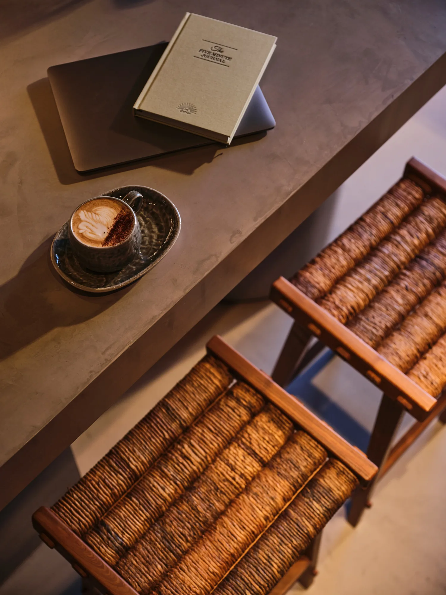 Cappuccino with latte art on a table beside books and woven wooden chairs in a bright, calm interior space.