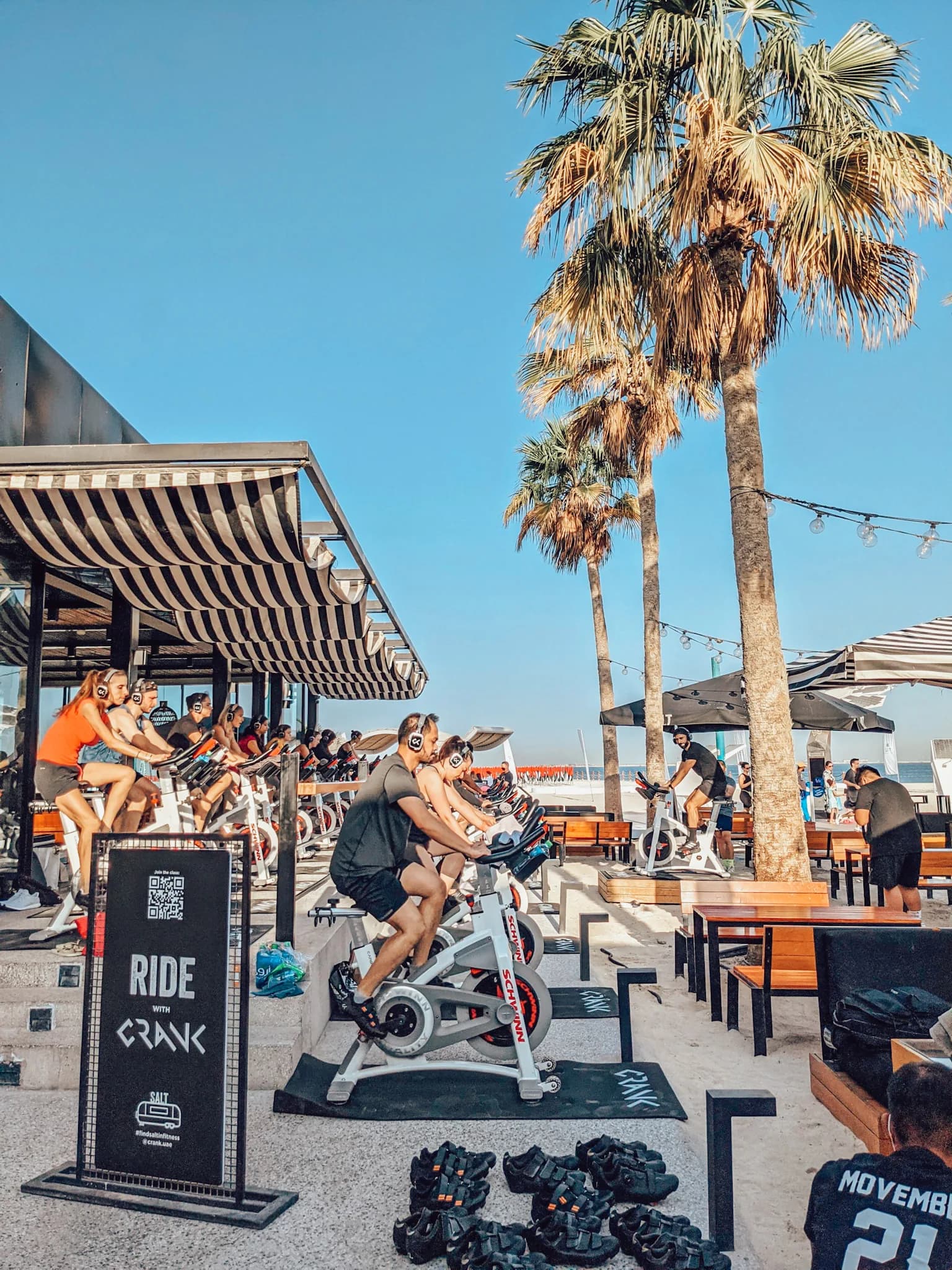 Outdoor spin class at beachside fitness studio with cyclists under canopies and palm trees
