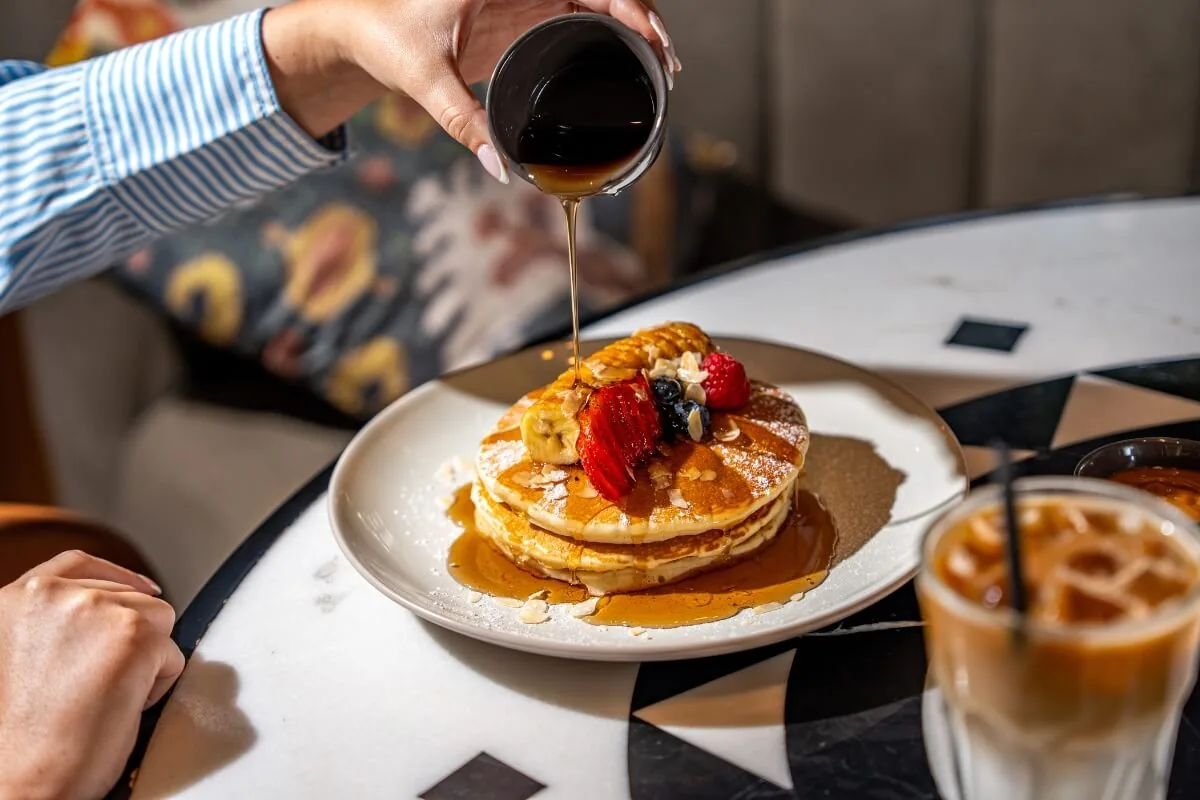 Maple syrup being drizzled over stacked pancakes with berries and powdered sugar