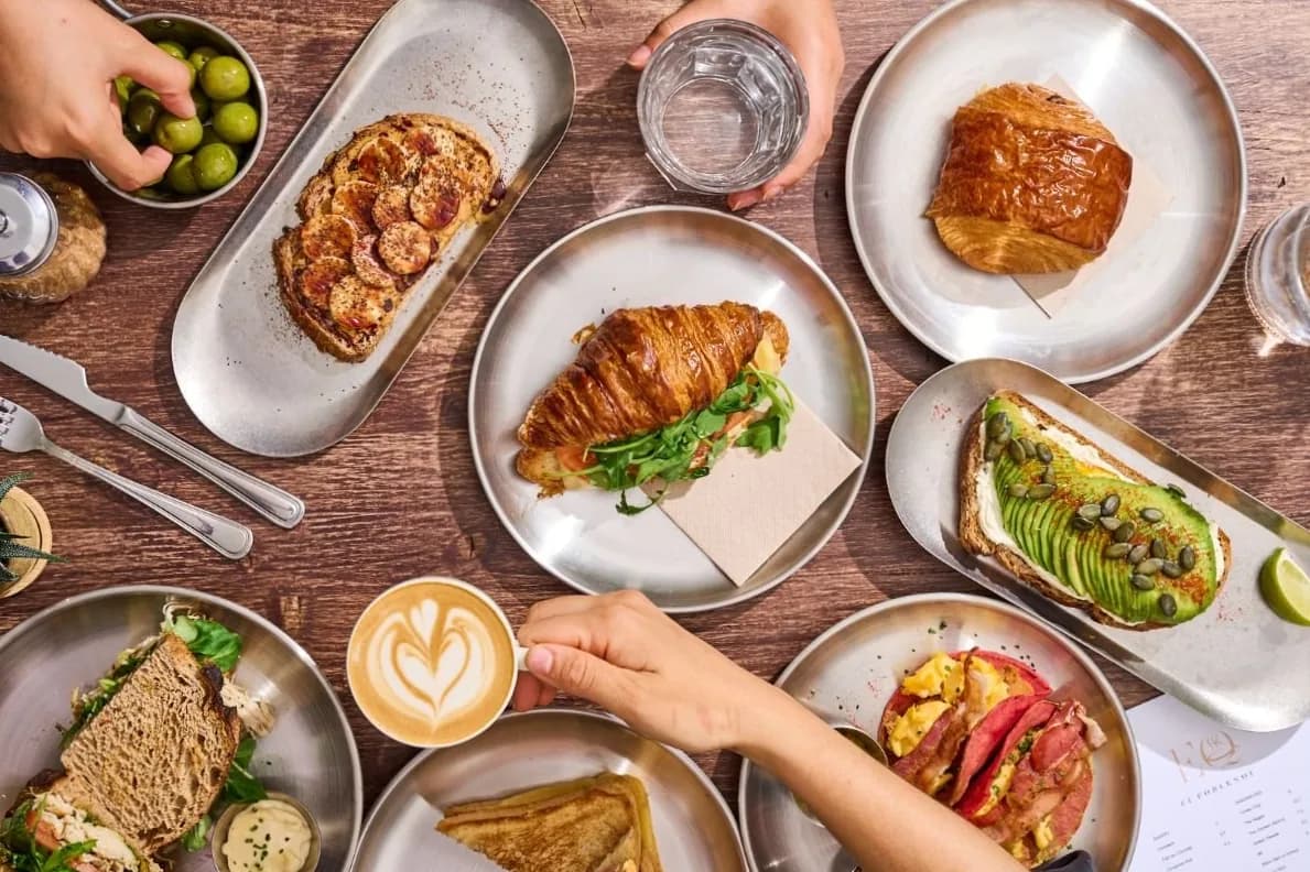 Overhead view of cafe breakfast dishes: croissants, toasts, coffee with latte art, and fresh ingredients on wooden surface