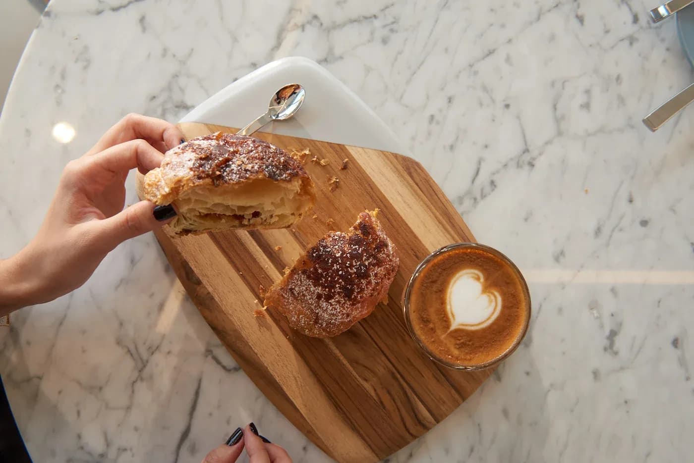 Hand holding sugar-dusted pastry with cappuccino and latte art on marble table