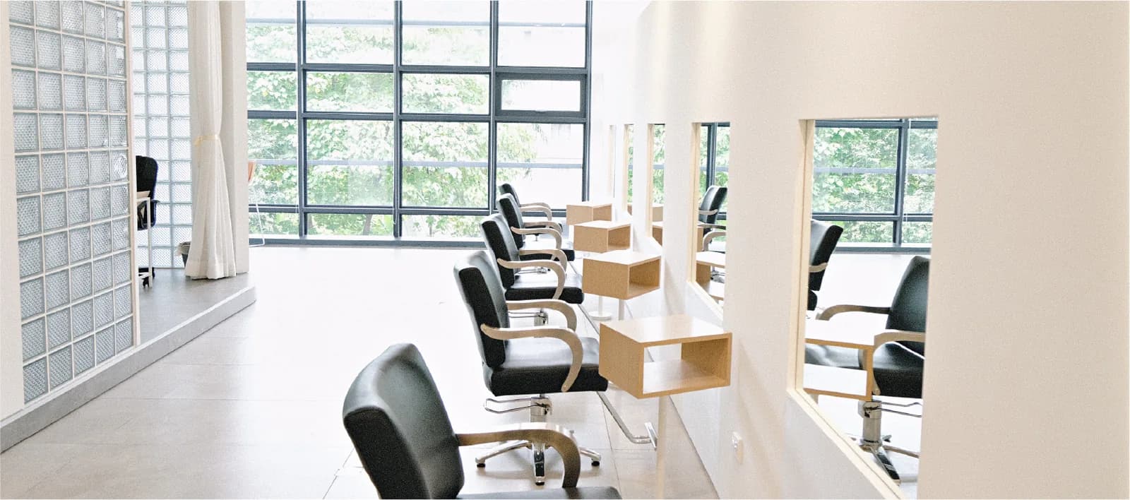 Modern hair salon with black styling chairs, wooden stations, and large windows with green views.