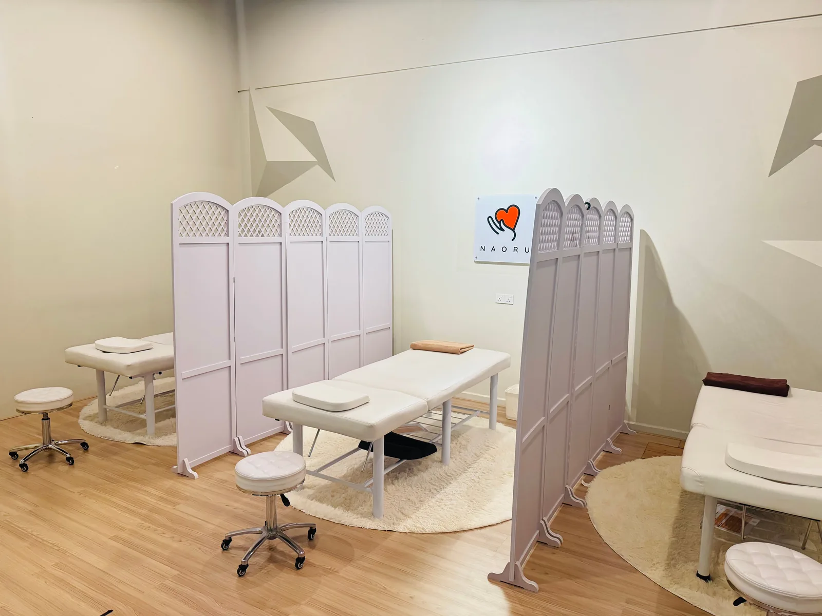 Spa treatment room with white massage beds, pink and gray privacy partitions, and pale wood flooring.