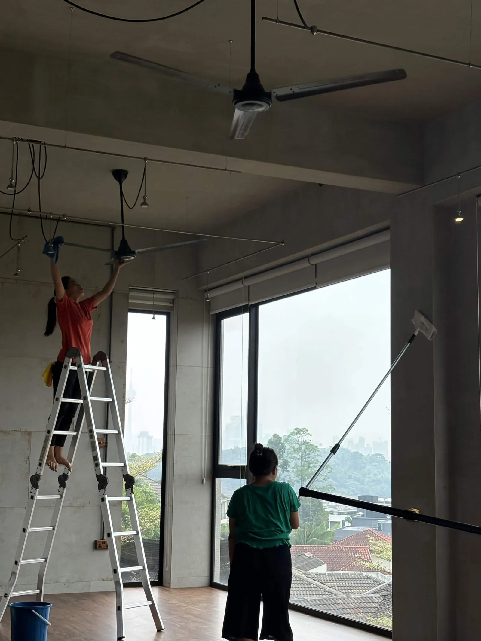 Technician on ladder installing ceiling fan while colleague assists in bright room with large windows