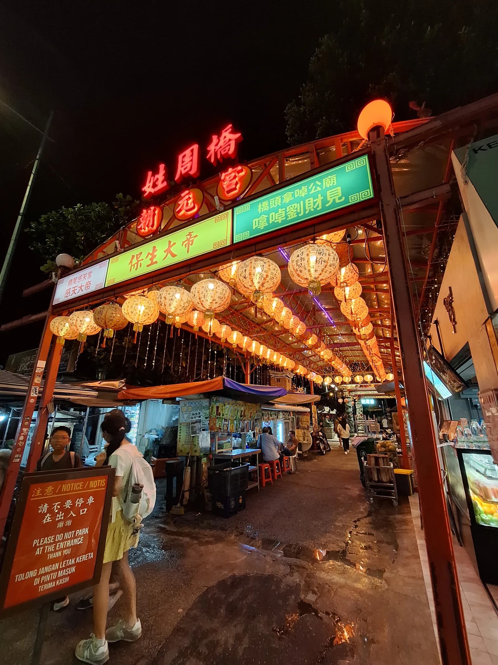 Night view of a Chinese food market entrance with neon signs and hanging golden lanterns, customers dining under covered walkway.