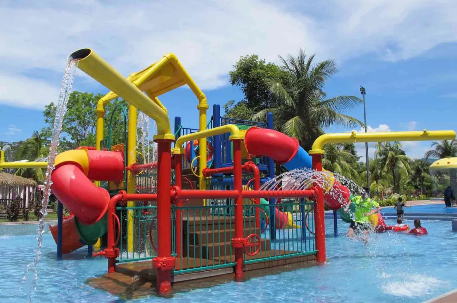 Bright red and blue children's water play structure with yellow slides in shallow pool, palm trees in background