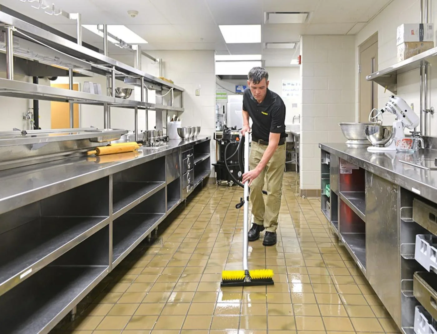 Worker cleaning commercial kitchen floor with push broom among stainless steel prep counters and equipment