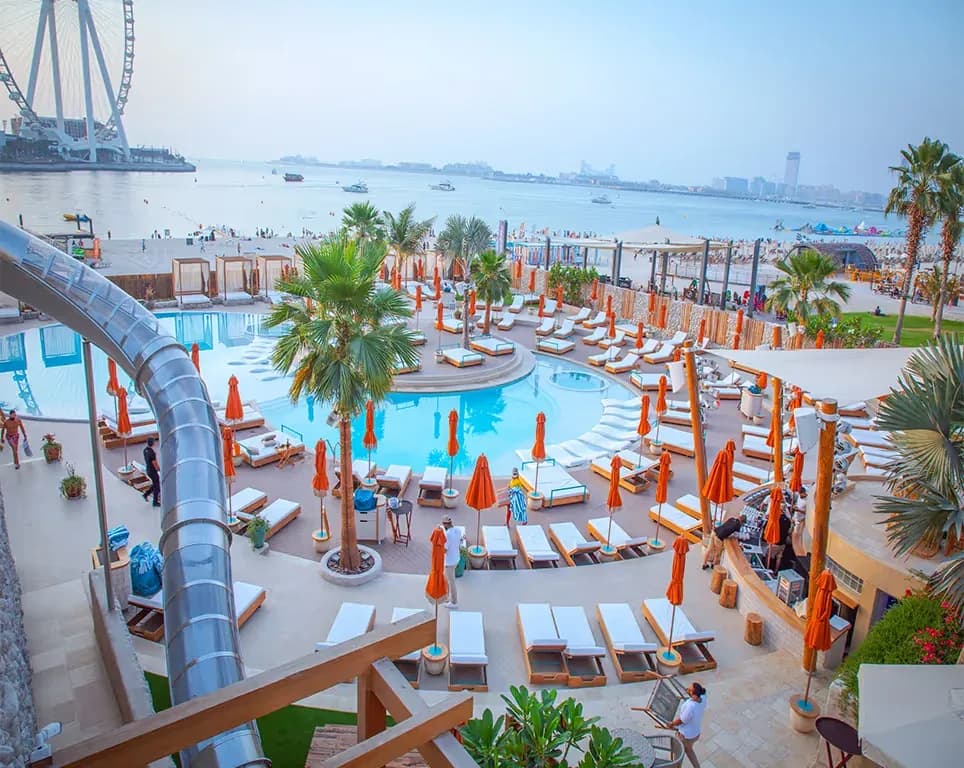 Resort pool area with white loungers, orange umbrellas, and beachfront beyond