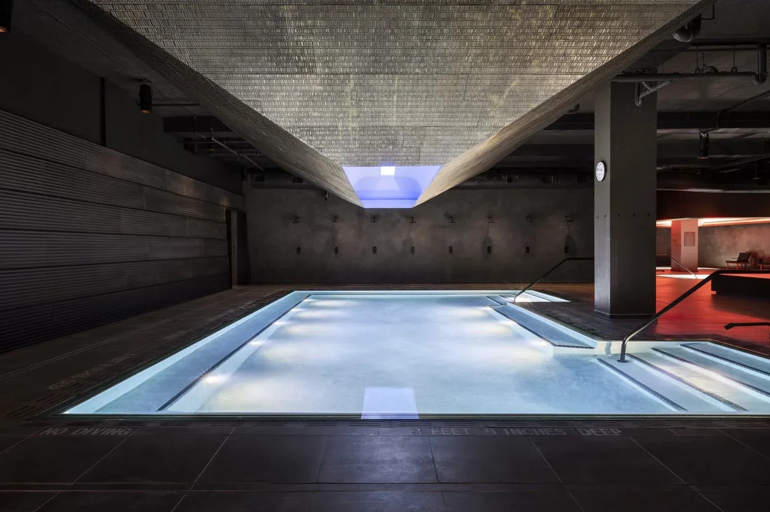 Modern indoor swimming pool with blue-lit water, concrete walls, and natural skylight in spa facility