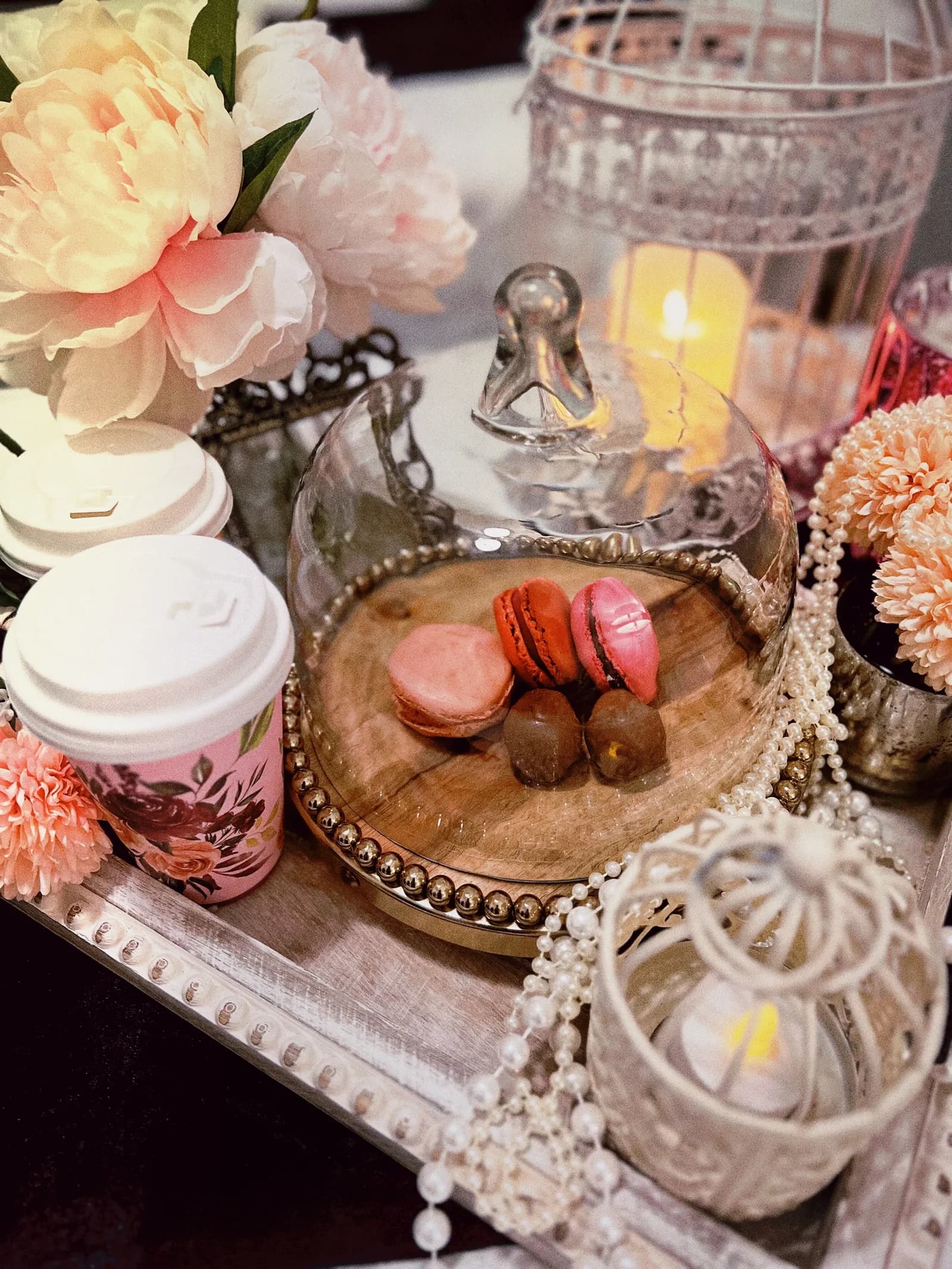 Colorful macarons and pastries displayed with pink peonies, candles, and pearls on wooden plate