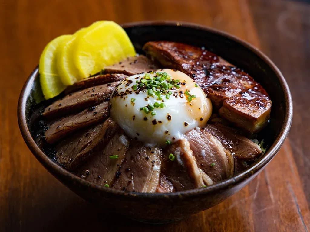 Bowl of meat donburi with soft-boiled egg, green onions, and pickled vegetables on wooden table