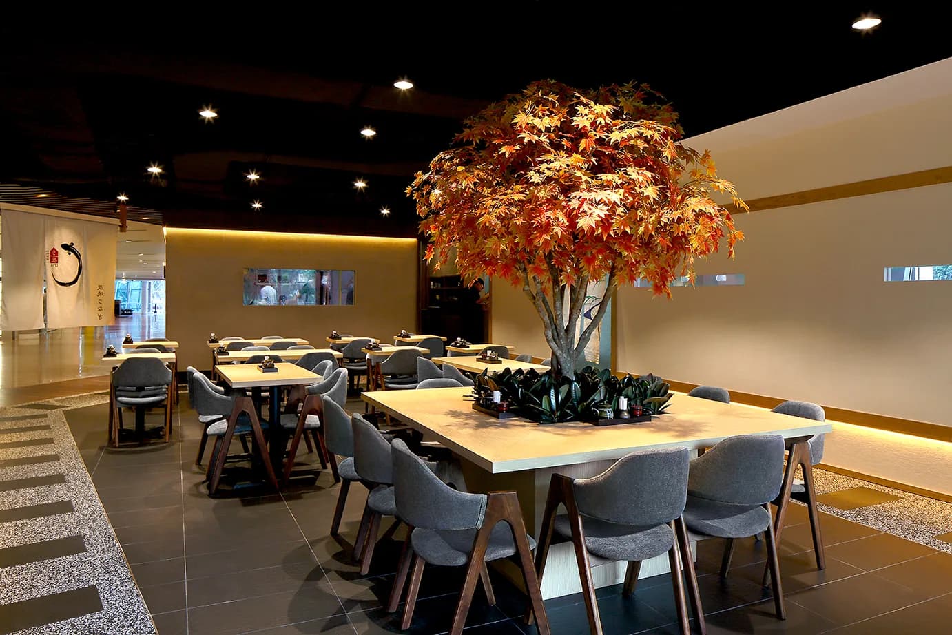Modern restaurant interior with dining tables, comfortable seating, and a large decorative maple tree under warm lighting