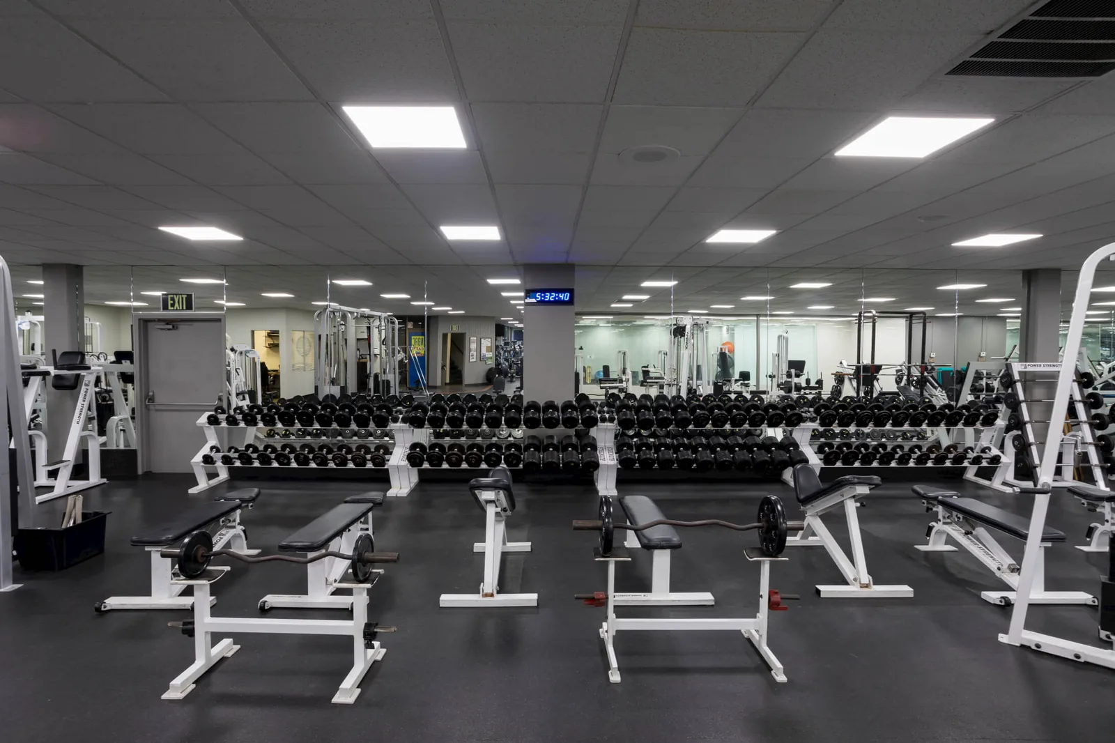 Gym floor with rows of dumbbells, weight benches, and cardio machines under bright ceiling lights