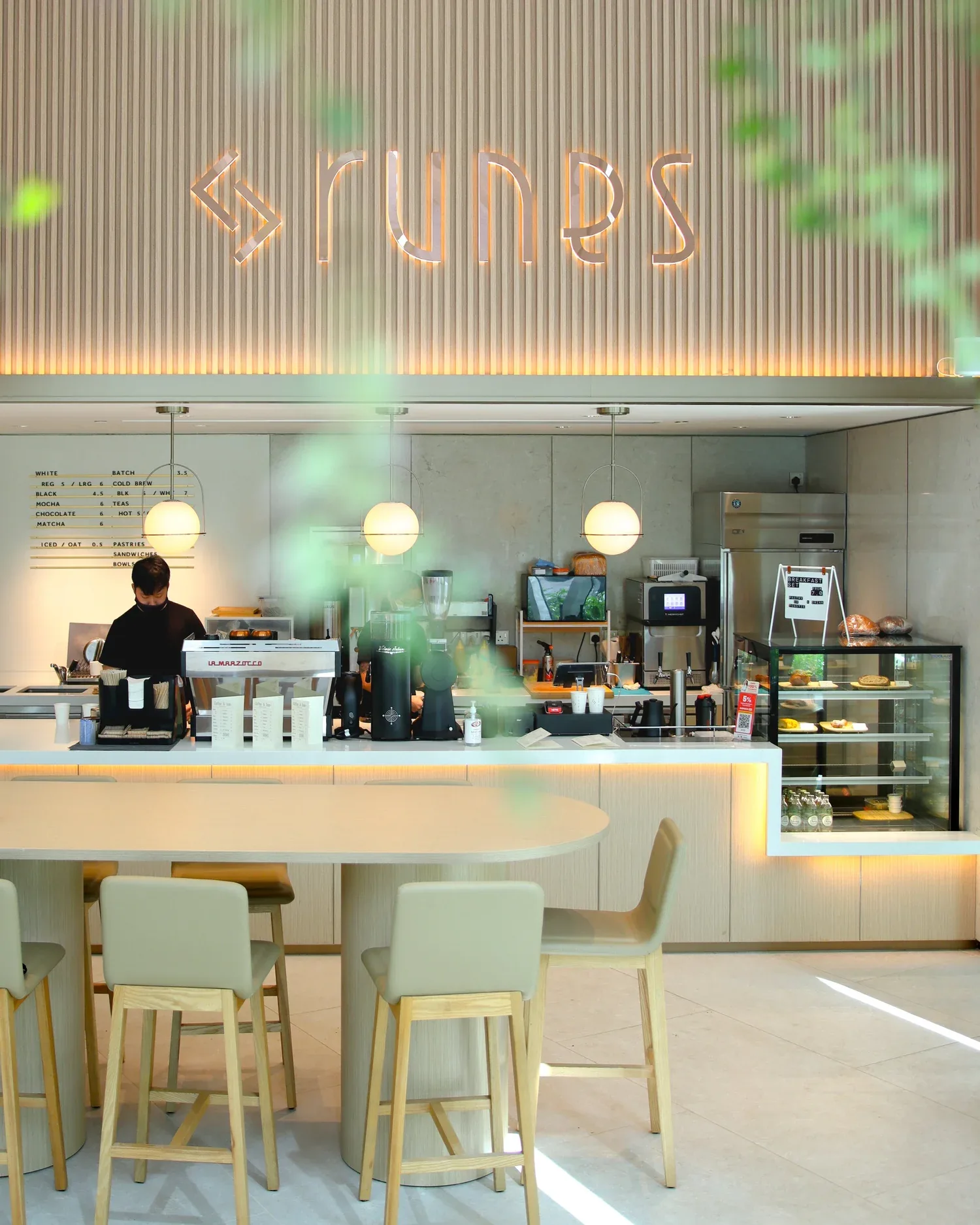 Barista working at counter in modern café with neon 'GLYPH'S' sign and pastry display