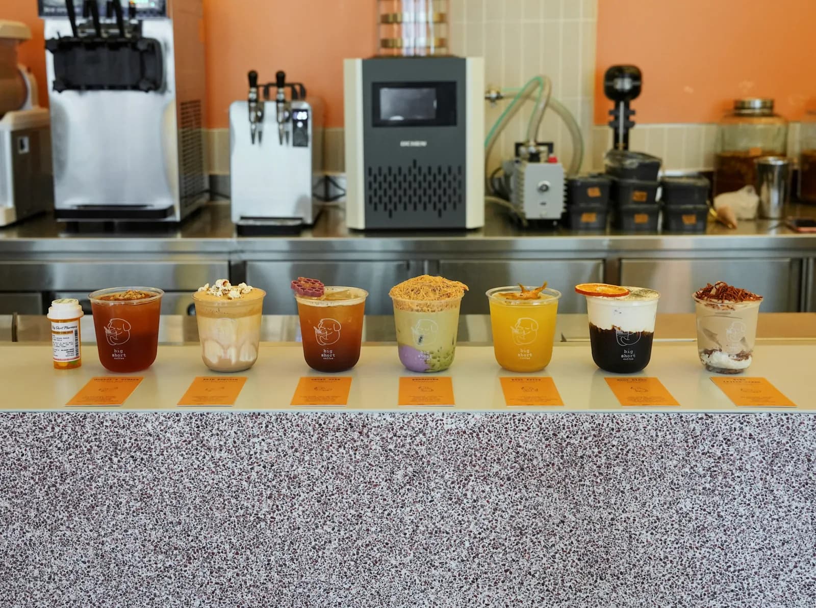 Seven specialty drinks lined up on a café counter with orange walls and equipment behind