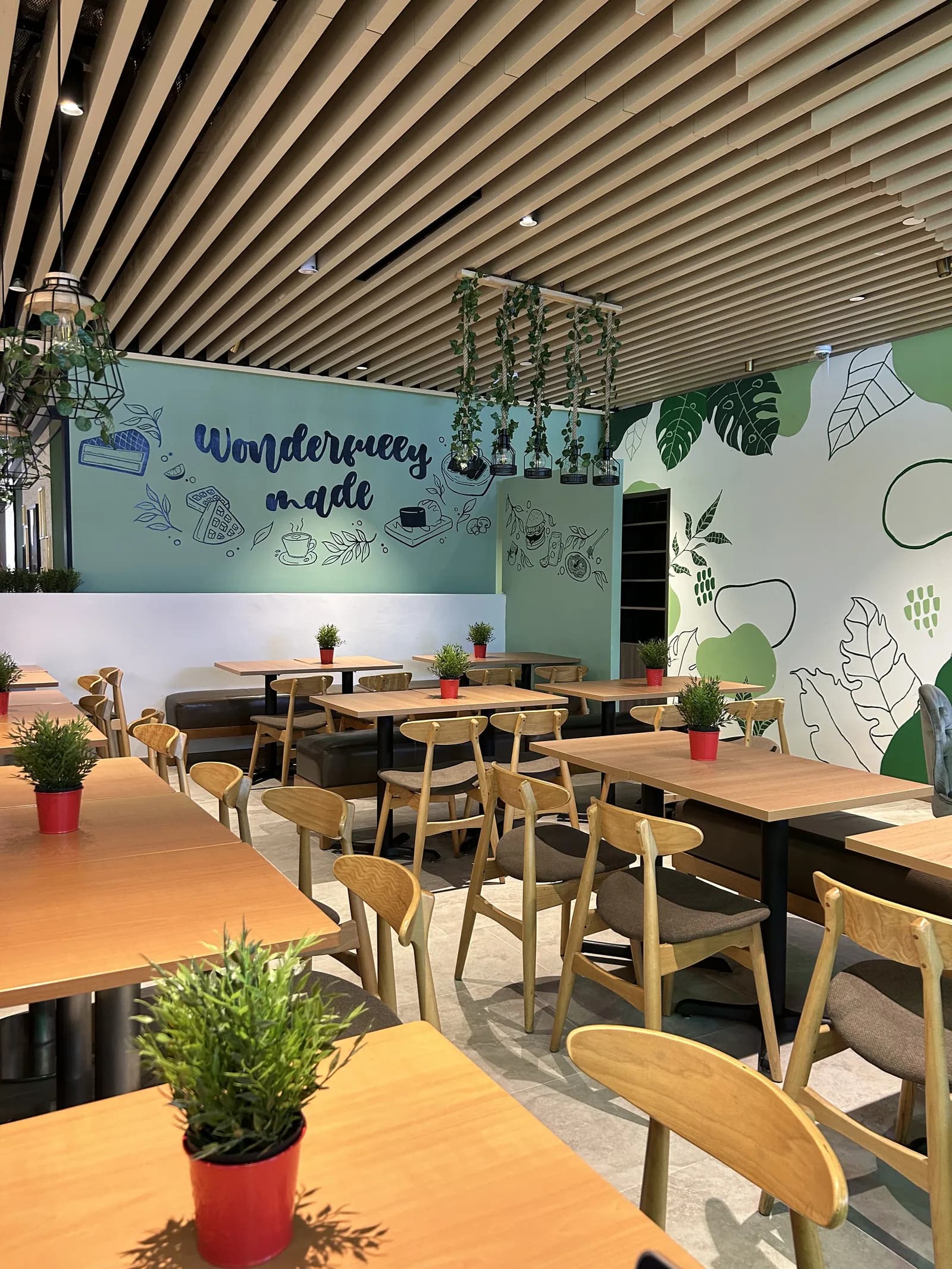 Modern café interior with wooden furniture, green wall murals, hanging plants, and contemporary ceiling design