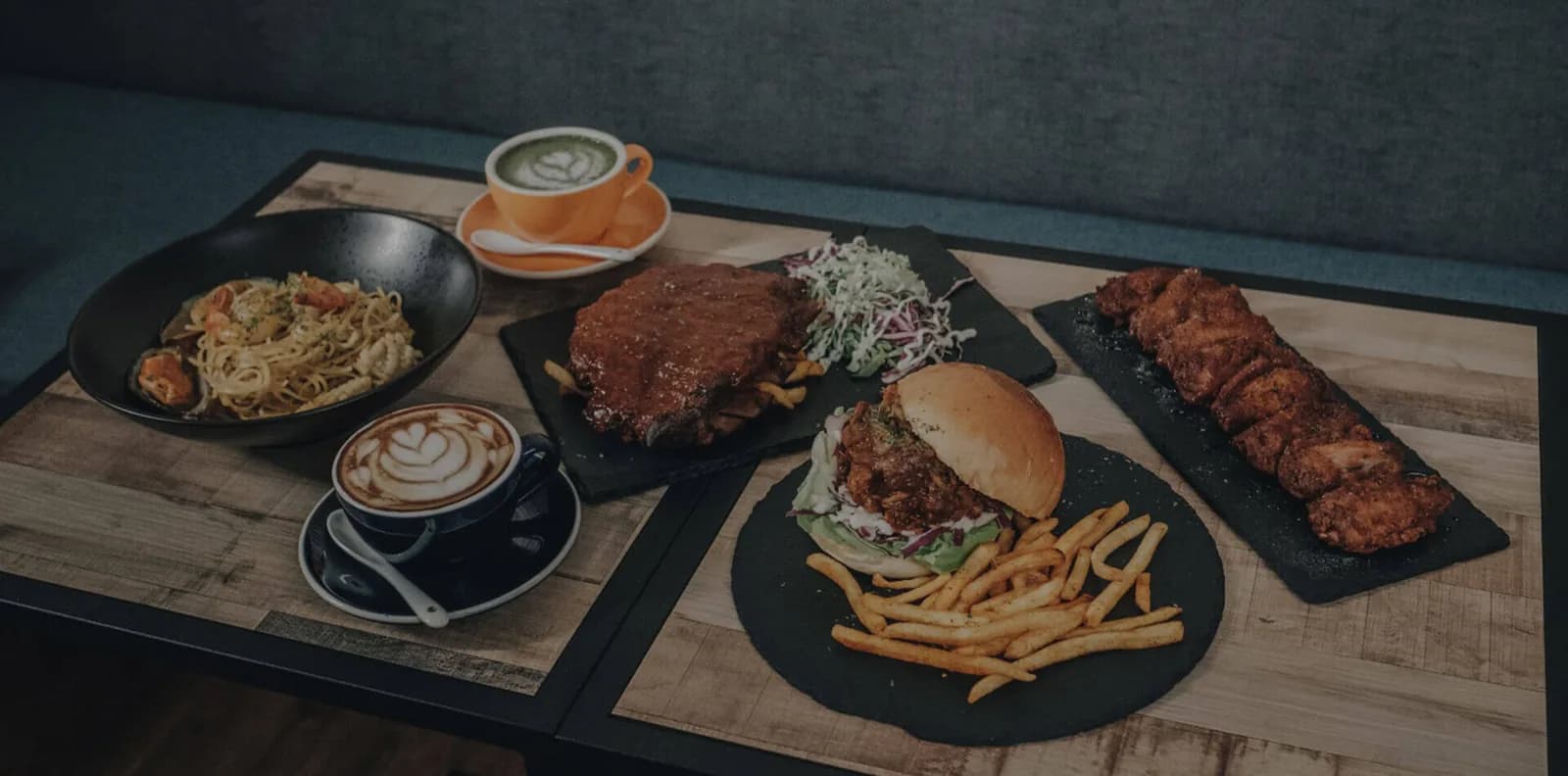 Assorted cafe dishes including noodles, fried chicken burger, steak, and specialty coffees arranged on wooden serving boards