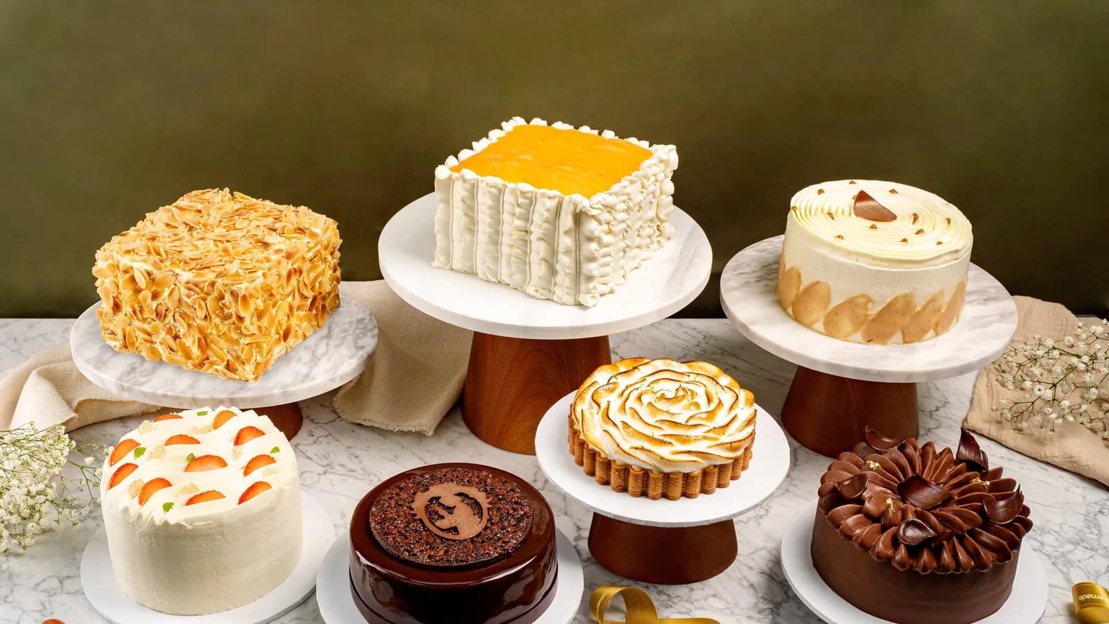 Six decorated cakes displayed on white plates and cake stands on a marble surface