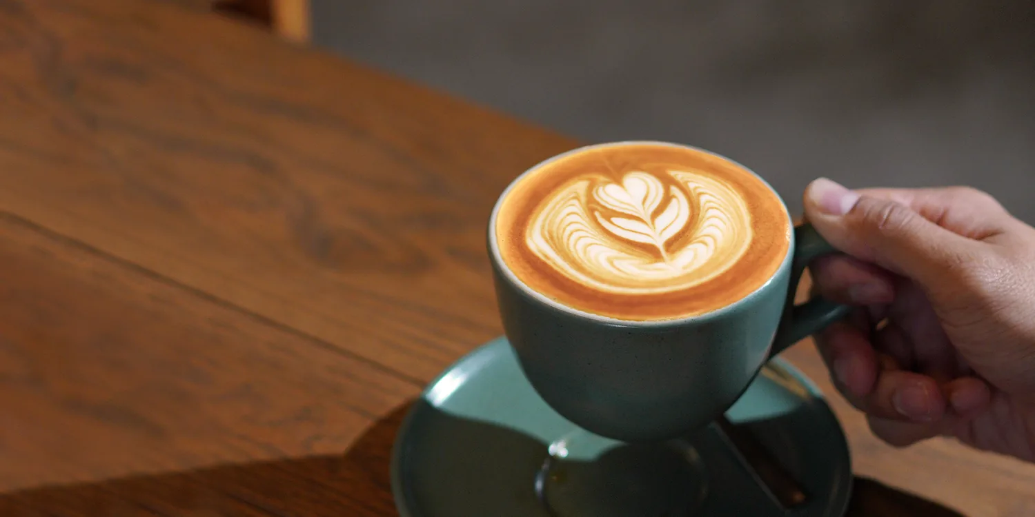 Hand holding teal cup of coffee with latte art leaf design on wooden table