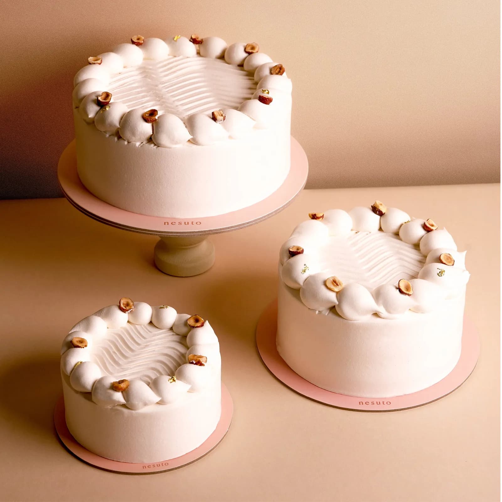 Three white cakes with cream piping and gold leaf decorations on pink bases