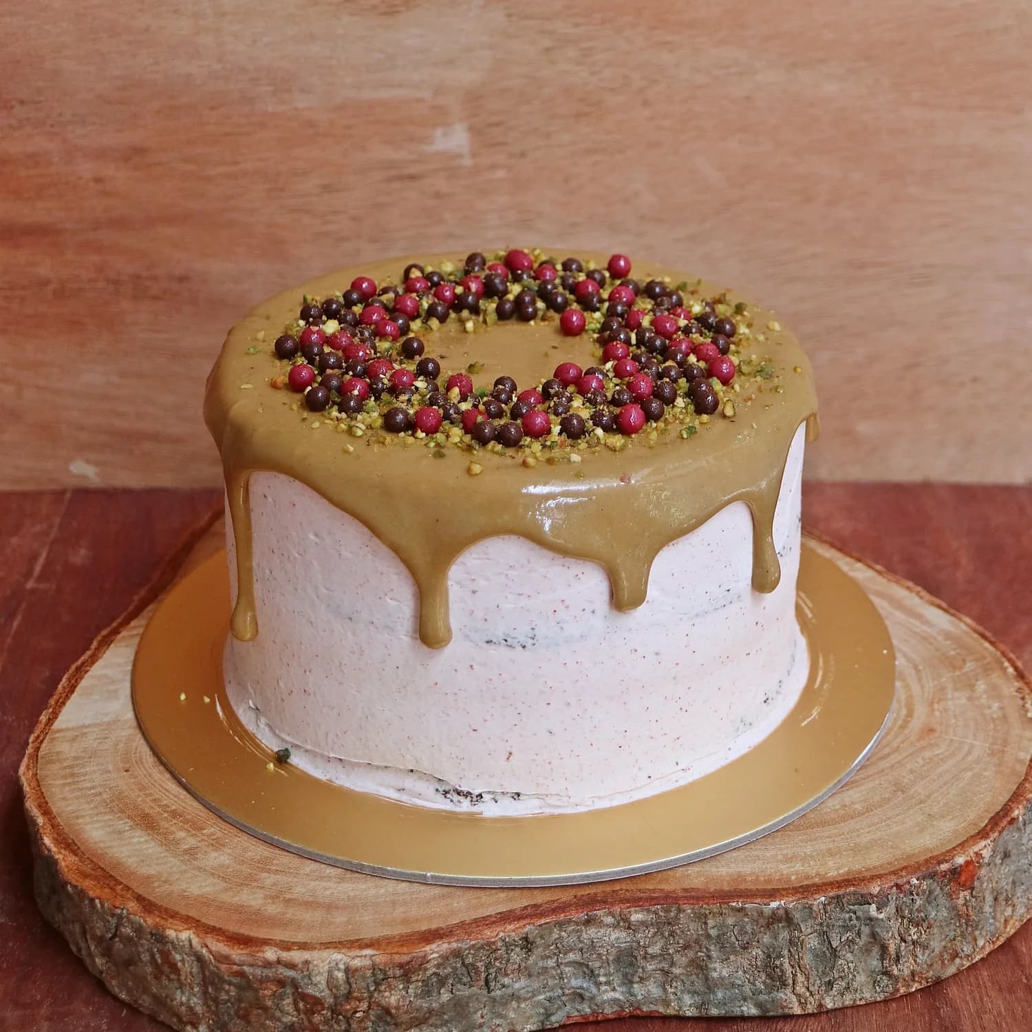 Layer cake with pink frosting, caramel drip, pistachios, and red berries on wooden board