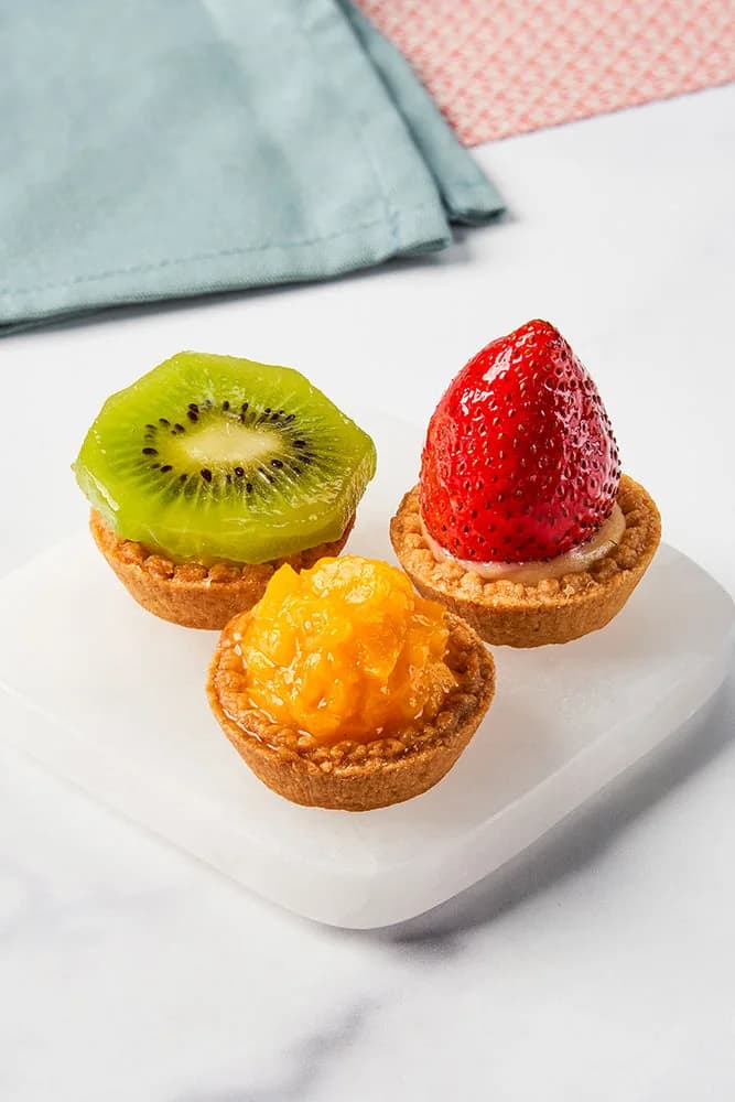 Three colorful fruit tarts with kiwi, strawberry, and mango on white plate