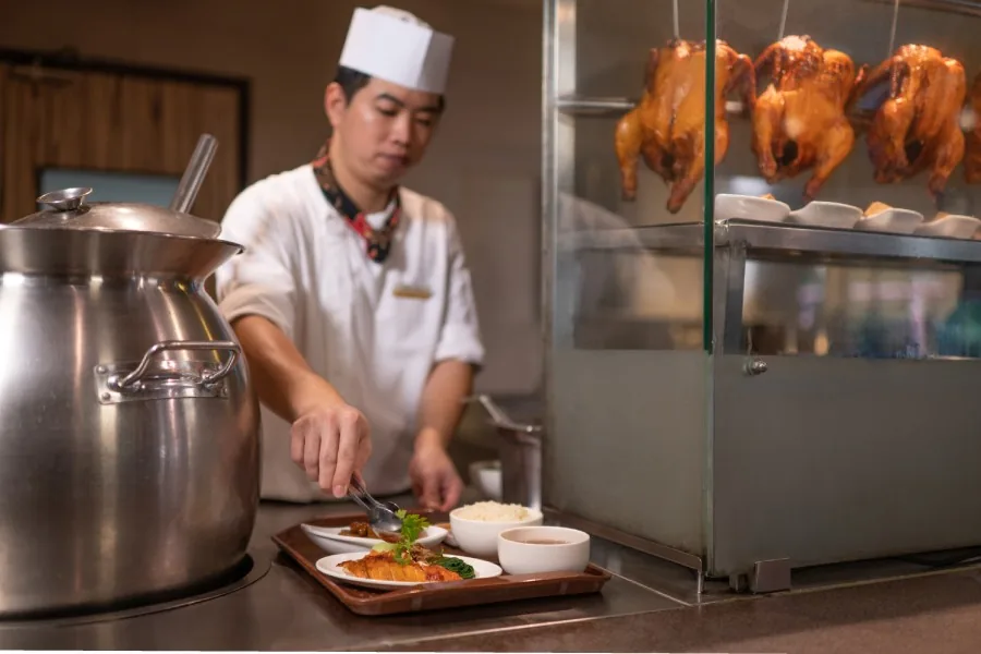Chef plating a dish in open kitchen with roasted ducks displayed in glass case