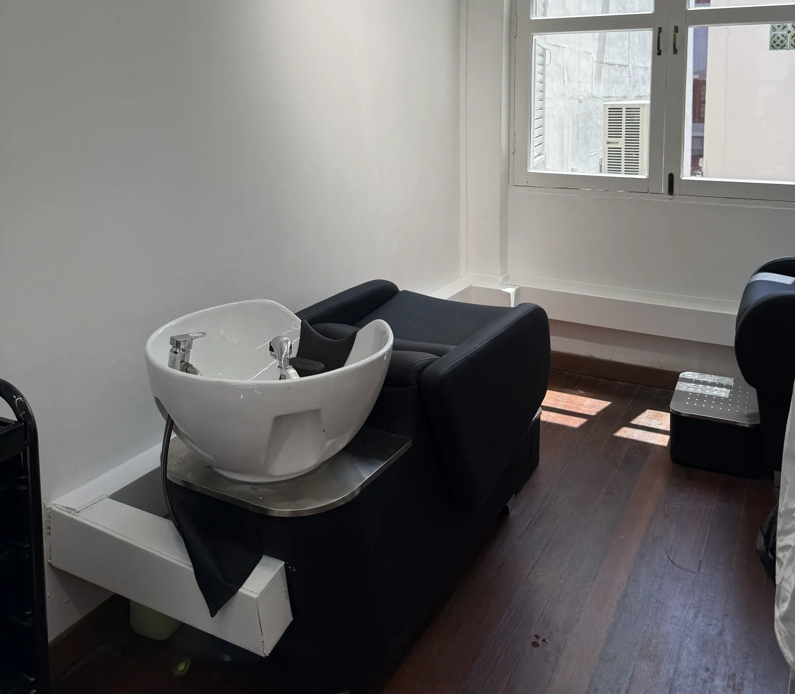 White bowl sink station with black cushioned chair in bright, modern salon interior with windows