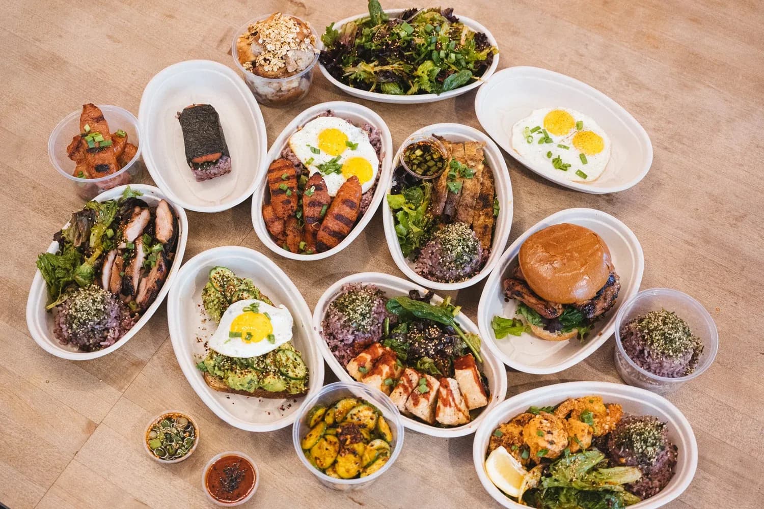 Overhead view of multiple plated dishes including grain bowls, eggs, grilled meats, burger, and fresh greens