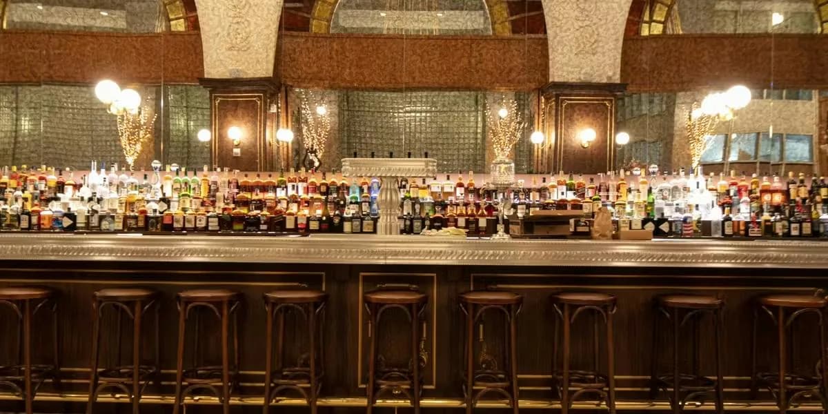 Well-stocked bar with numerous bottles illuminated by pendant lights, wood paneling and upholstered stools