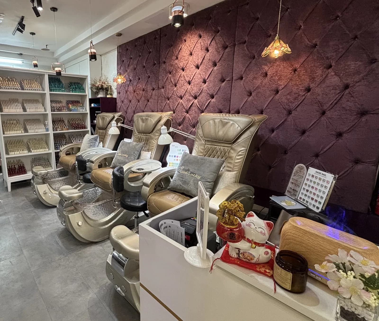 Modern nail salon interior with cream pedicure chairs, purple quilted wall, pendant lights, and polish display shelves