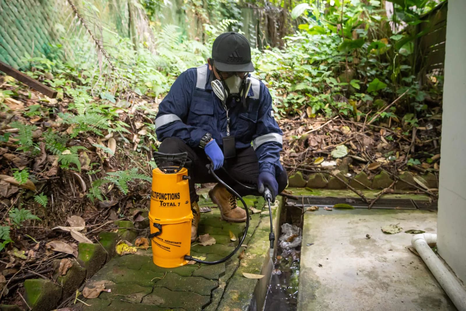 Pest control technician in blue uniform and protective gear working with treatment equipment among vegetation.