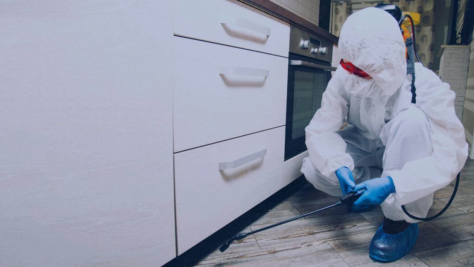 Pest control technician in protective suit spraying treatment along kitchen cabinet baseboard