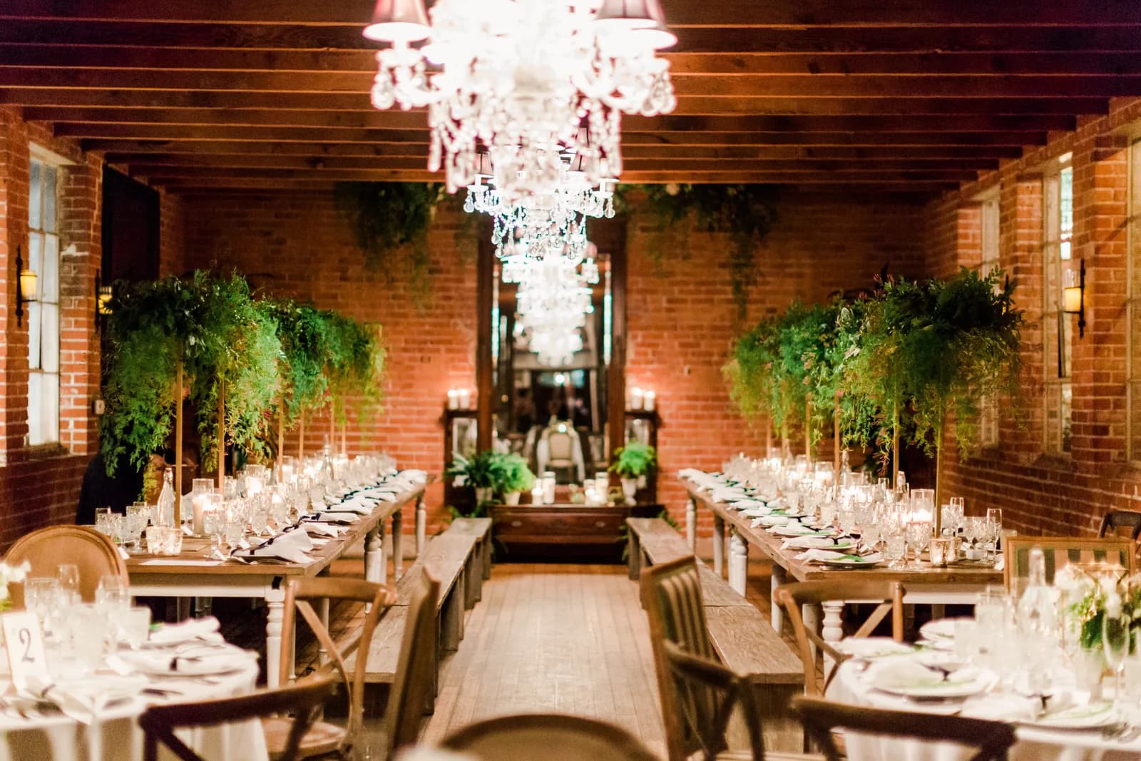 Upscale wedding reception room with brick walls, crystal chandeliers, long tables set for dining, and lush hanging plants