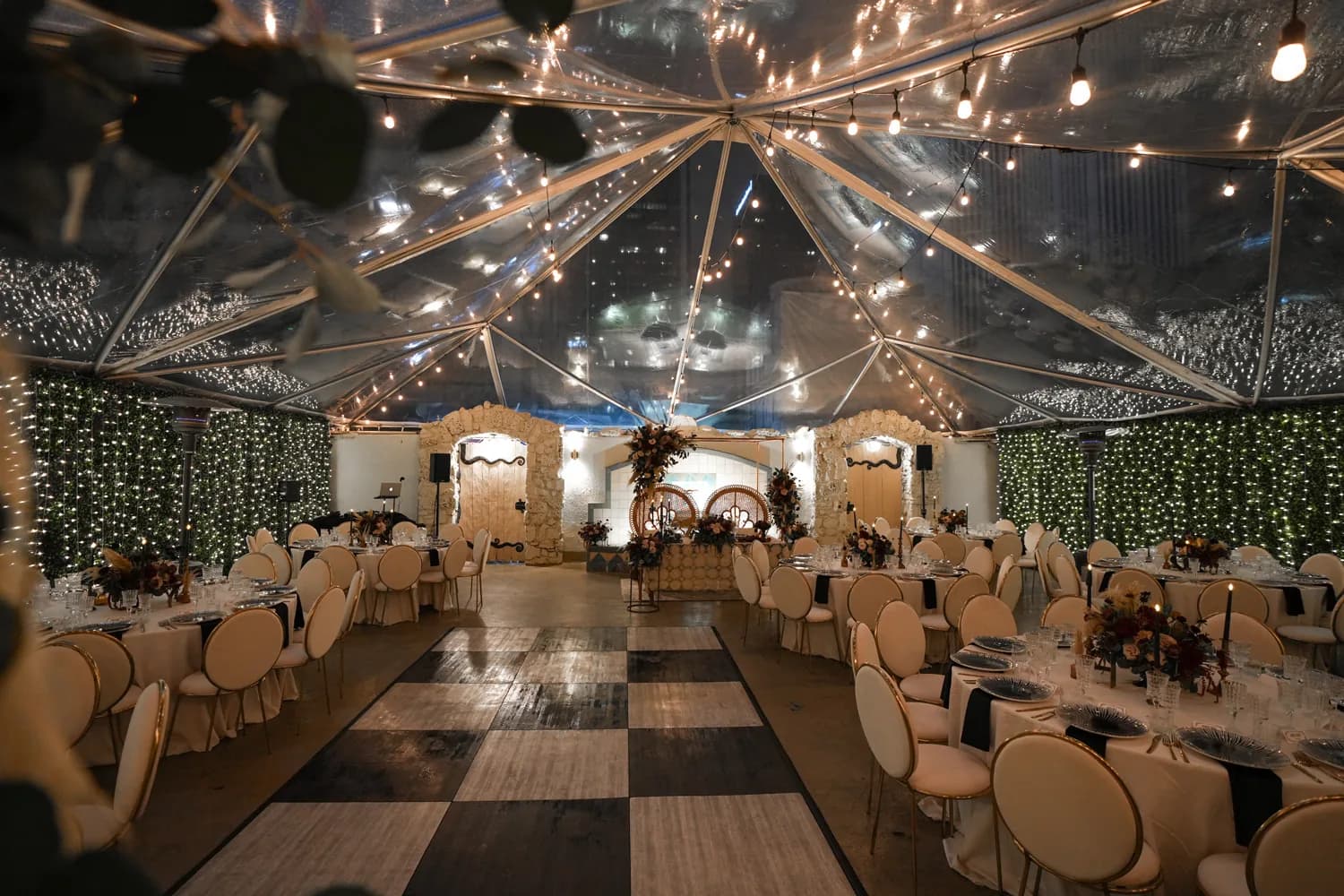 Elegant wedding venue with clear tent ceiling, warm string lights, decorated tables and gold chairs